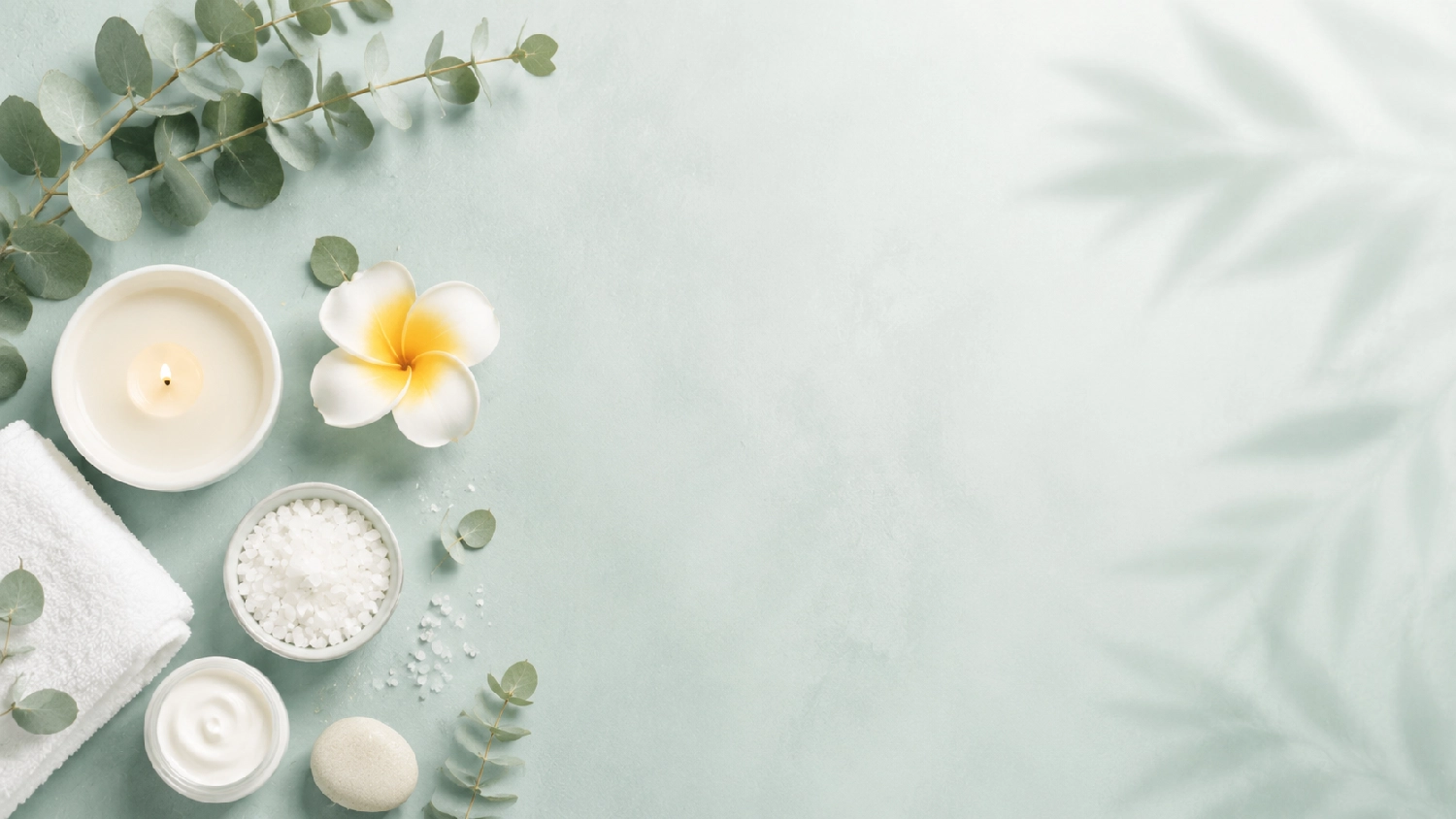 Marina Litwin - Behandlungen für Gesicht, Körper, Hände und Füße - Eine Draufsicht auf ein im Spa-Stil arrangiertes Arrangement vor hellgrünem Hintergrund, mit einem weißen Handtuch, Eukalyptuszweigen, Kerzen, Meersalz, Frangipaniblüten und einer Flasche Öl.
