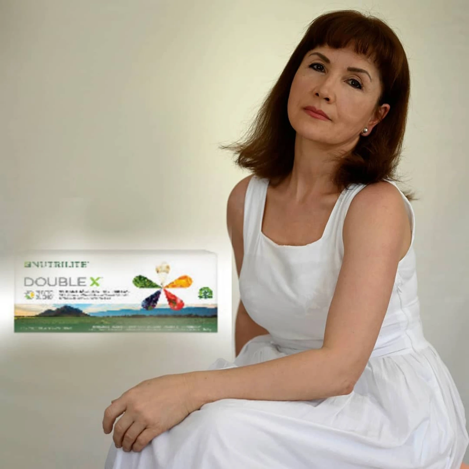 Marina Litwin - Behandlungen für Gesicht, Körper, Hände und Füße -  Eine ältere Frau mit schulterlangem braunem Haar und Pony sitzt in einem weißen ärmellosen Kleid und posiert neben einer Packung Nutrilite Double X Multivitaminpräparate.