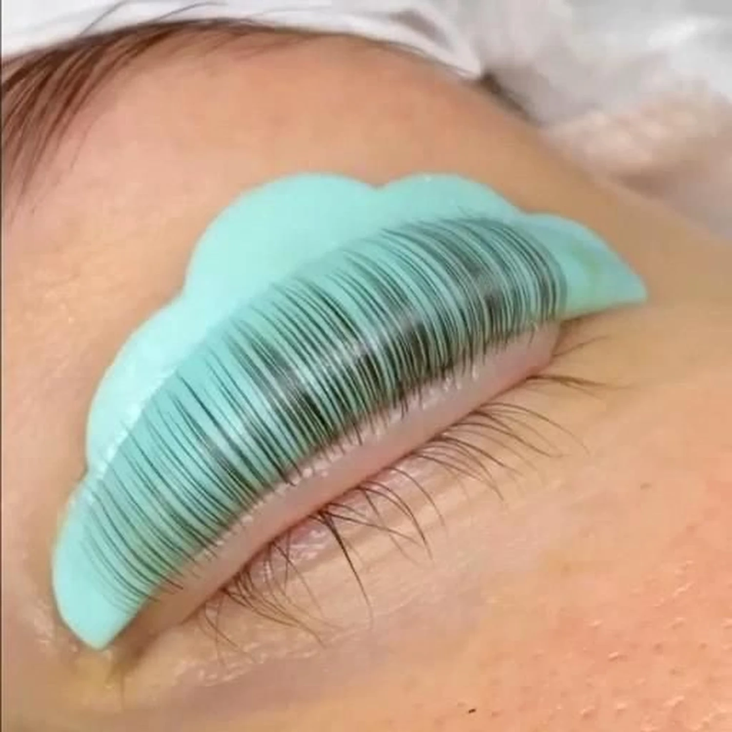 Marina Litwin - Behandlungen für Gesicht, Körper, Hände und Füße - Eine extreme Nahaufnahme des Auges einer Person, die sich einer Wimpernlifting-Behandlung unterzieht. Die natürlichen Wimpern werden sorgfältig gespreizt und auf eine hellblaue, gewellte Silikonabdeckung geklebt.