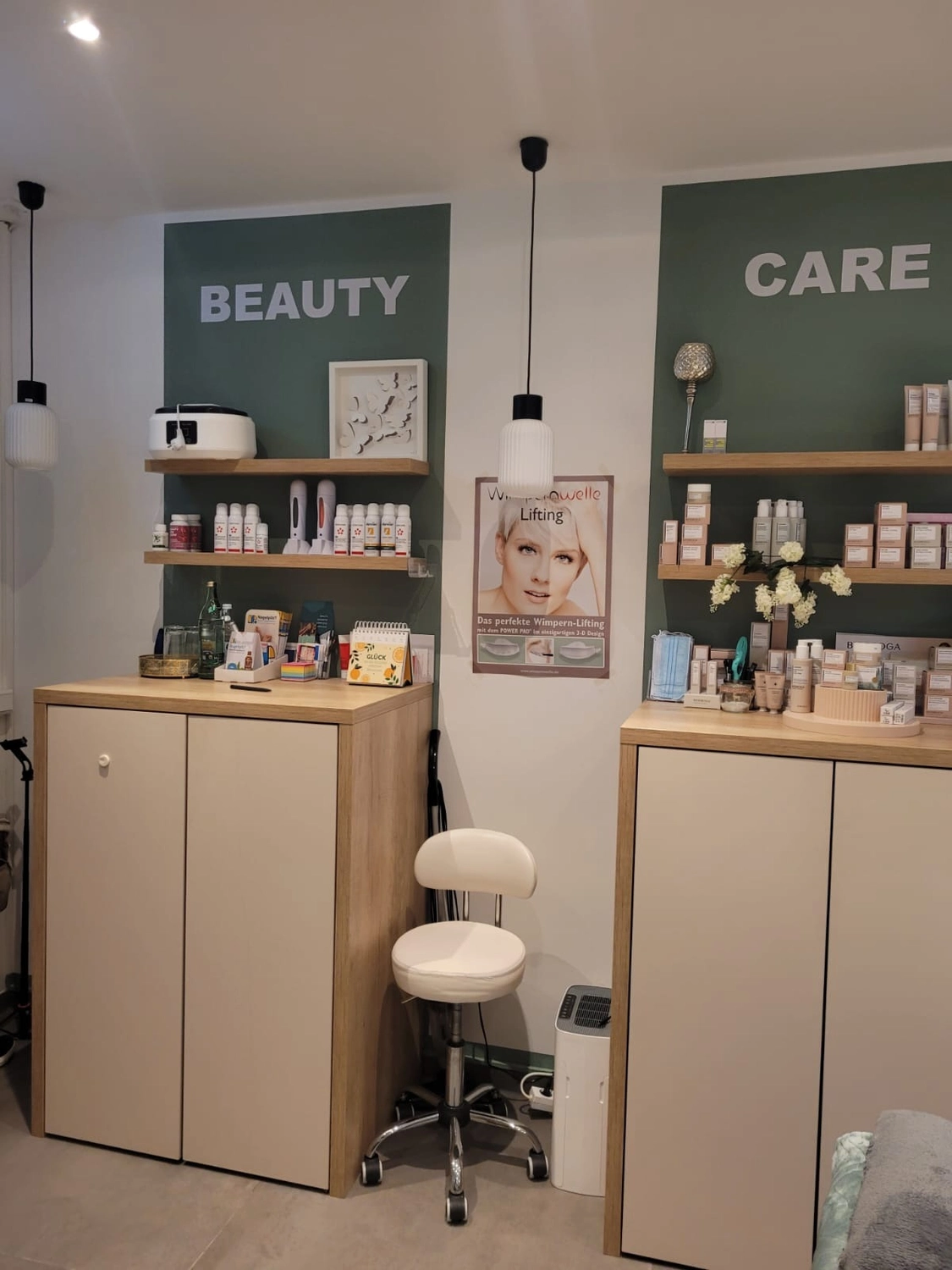 Marina Litwin - Behandlungen für Gesicht, Körper, Hände und Füße - Ein sauberes, modernes Saloninterieur mit zwei hellen Holzschränken vor weißen Wänden mit grünen rechteckigen Akzenten, die mit „BEAUTY“ und „CARE“ beschriftet sind. Regale präsentieren verschiedene Kosmetikprodukte und -werkzeuge, während ein weißer verstellbarer Hocker zwischen den Schränken unter minimalistischen Pendelleuchten steht.