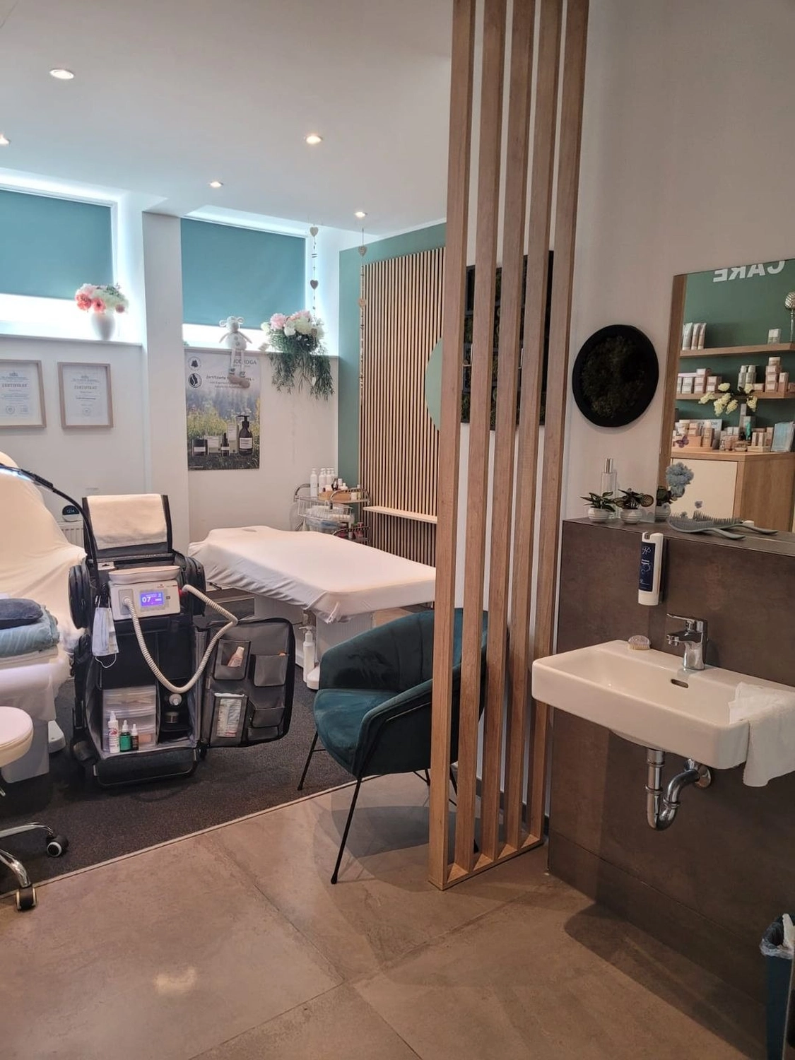 Marina Litwin - Behandlungen für Gesicht, Körper, Hände und Füße -  Ein modernes, gut beleuchtetes medizinisches Spa oder Behandlungsraum mit einem Gesichtsbehandlungsgerät, Behandlungsliegen, einem türkisfarbenen Samtsessel und einer minimalistischen Raumtrennung aus Holzlatten neben einem an der Wand montierten Waschbecken.