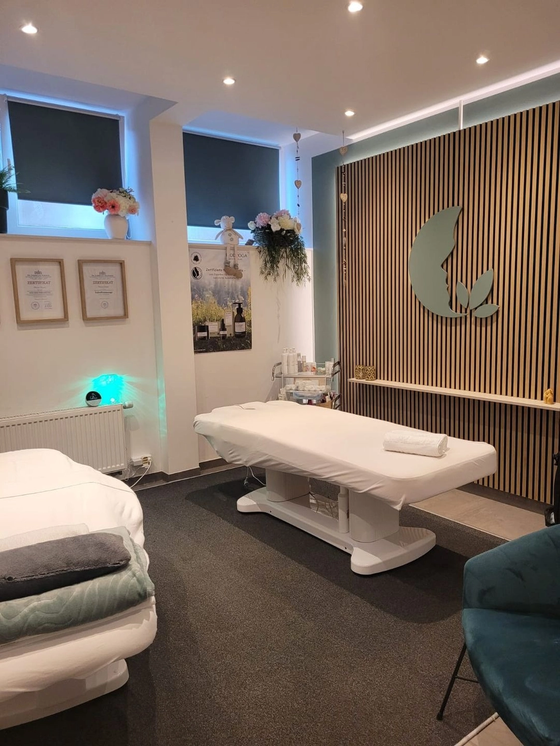Marina Litwin - Behandlungen für Gesicht, Körper, Hände und Füße - Ein moderner, minimalistischer Spa-Behandlungsraum mit einer weißen elektrischen Massageliege als zentralem Element. Die Rückwand ist mit vertikalen Holzlamellen und einem salbeigrünen Blattlogo verziert, während sanftes Einbaulicht eine ruhige, professionelle Atmosphäre schafft.