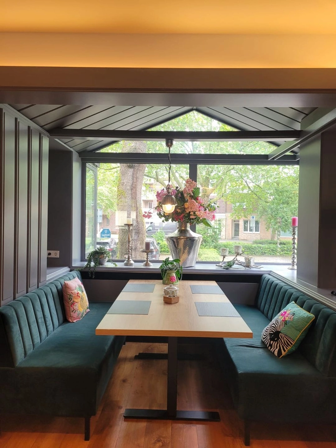 Marina Litwin - Behandlungen für Gesicht, Körper, Hände und Füße -  Eine moderne Essecke mit waldgrünen Samtsitzbänken um einen hellen Holztisch, die sich unter einem großen Fenster mit Blick auf üppig grüne Bäume befindet.