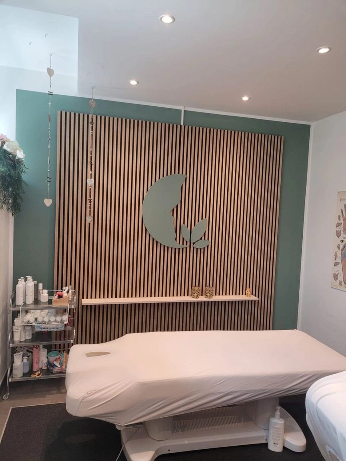 Marina Litwin - Behandlungen für Gesicht, Körper, Hände und Füße -  In einem modernen Behandlungsraum steht eine mit einem weißen Laken bedeckte Massageliege vor einer Wandverkleidung aus Holzlamellen mit einem salbeigrünen Blattlogo.