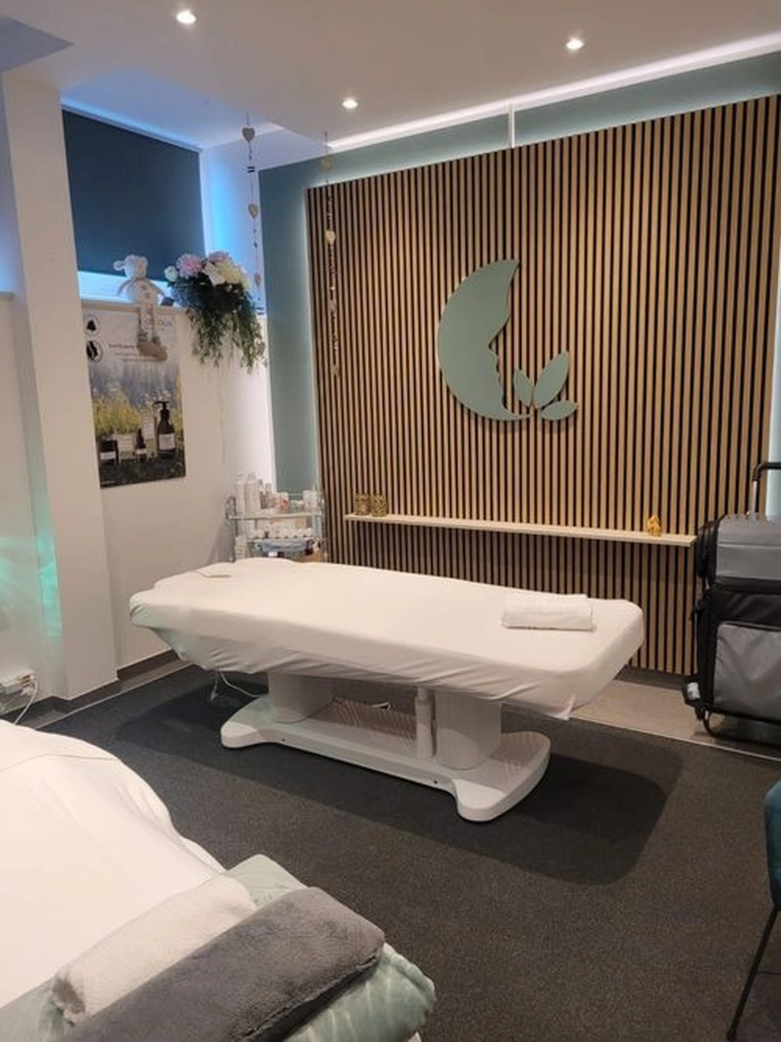 Marina Litwin - Behandlungen für Gesicht, Körper, Hände und Füße - Ein moderner Spa-Behandlungsraum mit einer weißen elektrischen Massageliege, einer Akzentwand aus Holzlatten mit einem Blattlogo und sanfter Einbaubeleuchtung.
