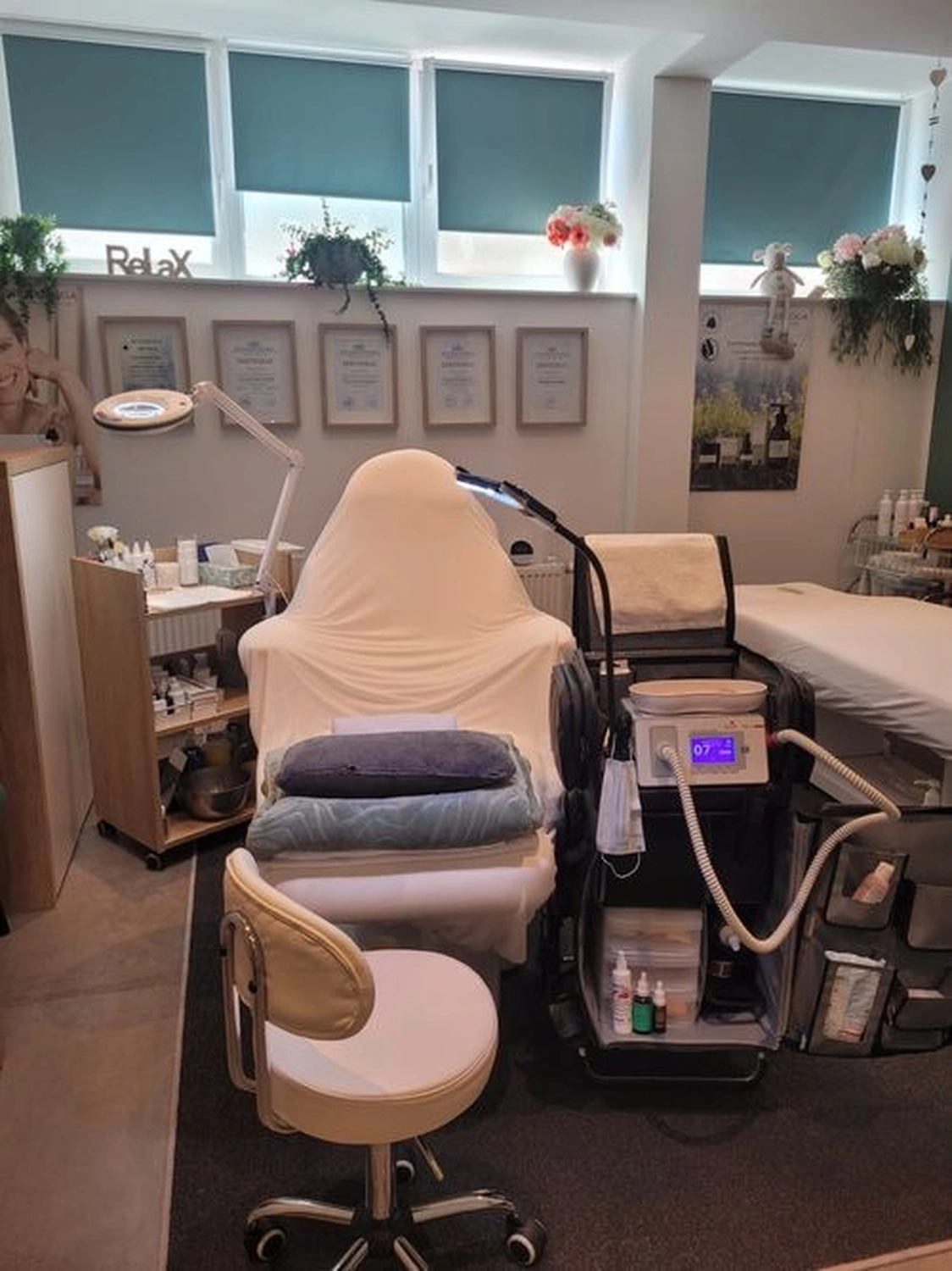 Marina Litwin - Behandlungen für Gesicht, Körper, Hände und Füße -  Ein Behandlungsraum in einem Spa oder einer ästhetischen Klinik mit einem mit weißem Stoff bezogenen Liegesessel, professionellen Hautpflegegeräten auf einem Wagen, einem Rollhocker und mehreren gerahmten Zertifikaten an der Wand.