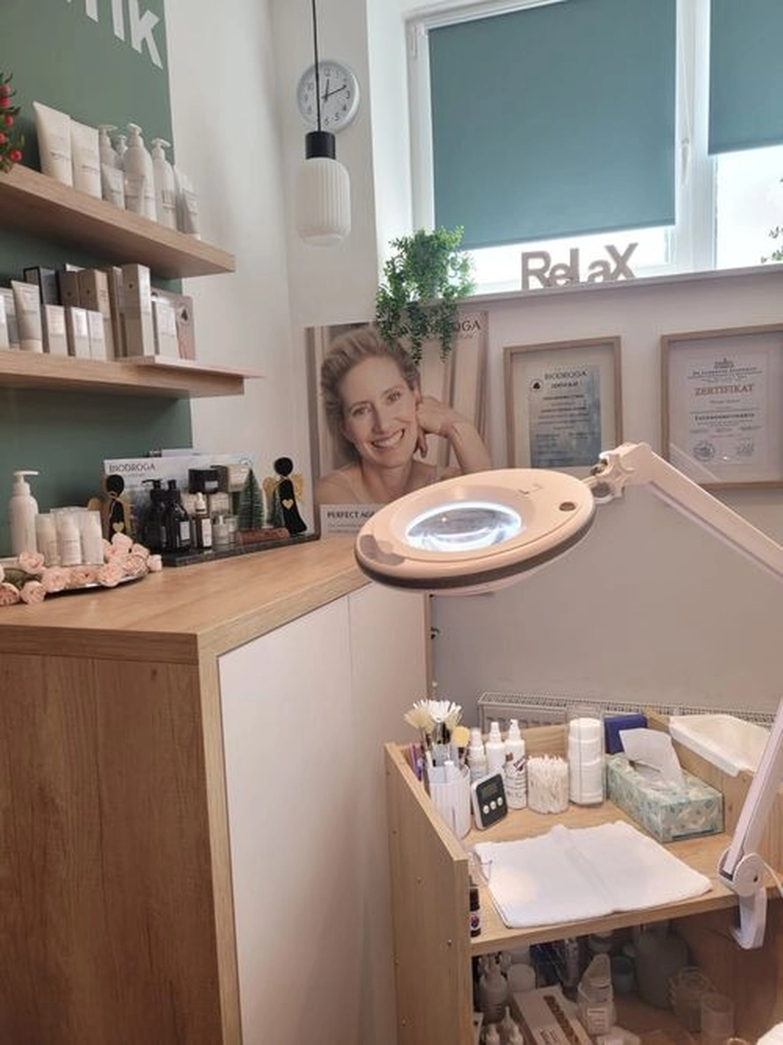 Marina Litwin - Behandlungen für Gesicht, Körper, Hände und Füße -  Eine hell erleuchtete, moderne Hautpflegeklinik oder ein Schönheitssalon mit Holzregalen, die mit professionellen Produkten bestückt sind, einer Vergrößerungslampe über einem Servierwagen und gerahmten Zertifikaten an der Wand.