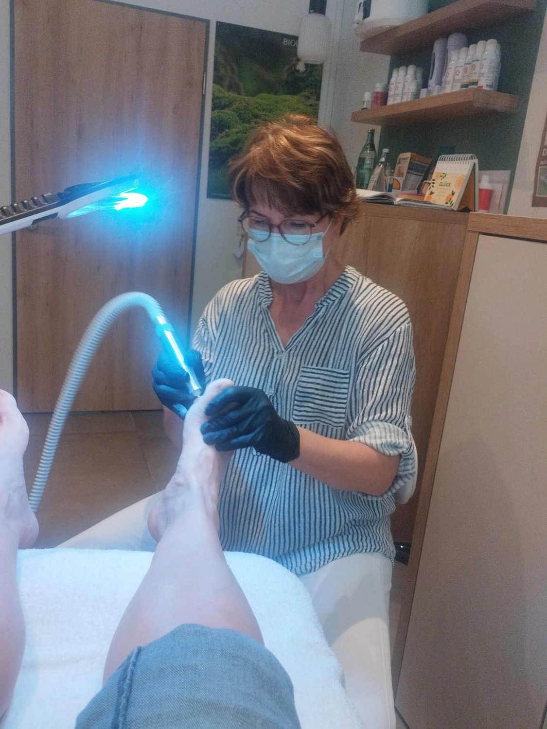 Marina Litwin - Behandlungen für Gesicht, Körper, Hände und Füße - Eine medizinische Fachkraft, die eine Gesichtsmaske und Handschuhe trägt, wendet ein Blaulichttherapiegerät am Fuß eines Patienten an.